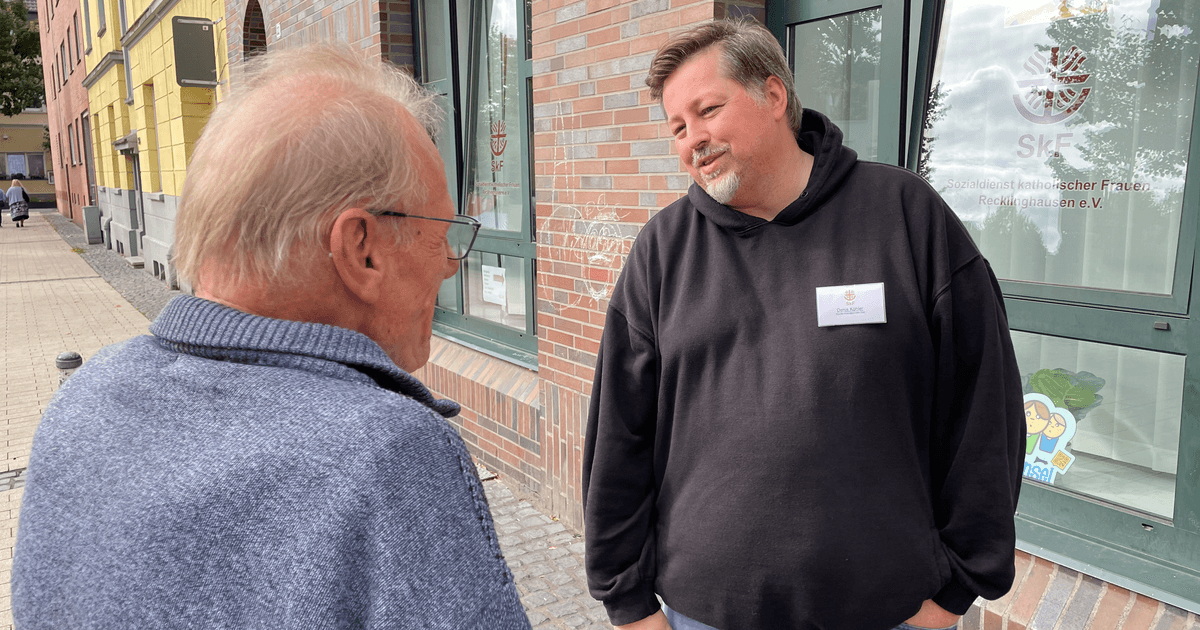 Ein Mann mit schwarzem Pullover spricht mit einem ihm gegenüberstehenden Mann