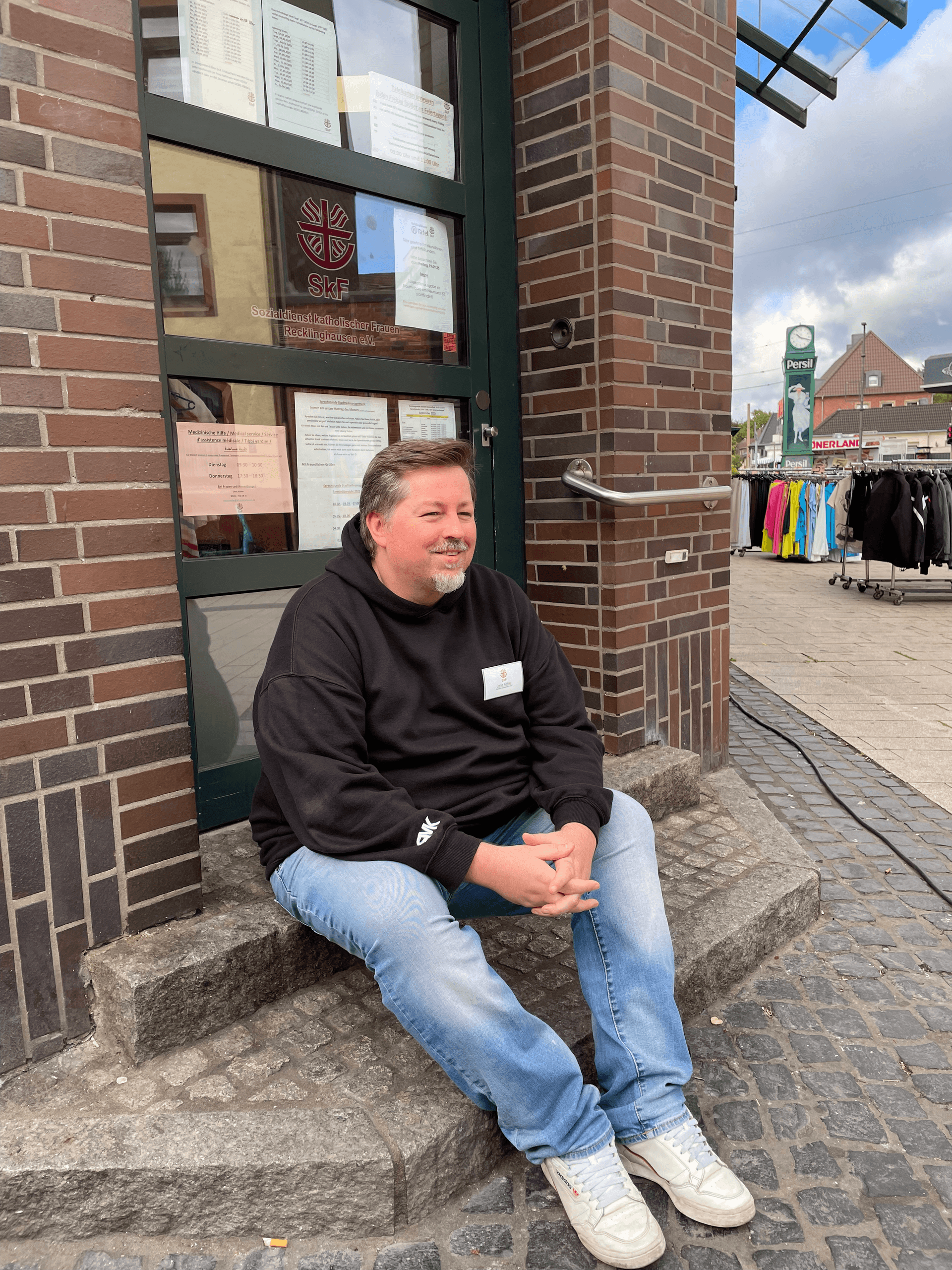 Der Stadtteilmanager des SkF Recklinghausen sitzt mit schwarzen Pullover vor dem Stadtteiltreff auf den Stufen und lächelt