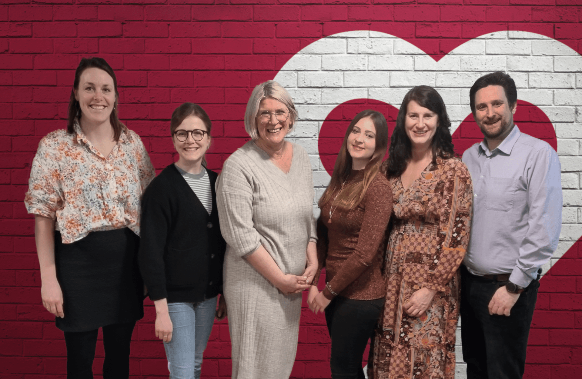 Das Team der Lotsentätigkeit steht lächelnd vor einer roten Backsteinwand mit Herzsymbol – ein engagiertes Team, das Unterstützung und Hilfe anbietet.