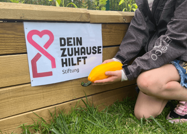 Kind erntet eine gelbe Zucchini aus einem Hochbeet mit dem Logo „Dein Zuhause hilft“.