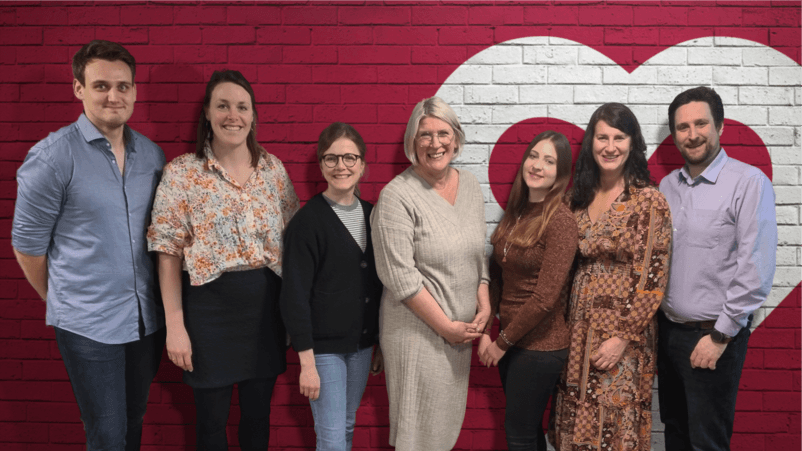 Das Team der Lotsentätigkeit steht lächelnd vor einer roten Backsteinwand mit Herzsymbol – ein engagiertes Team, das Unterstützung und Hilfe anbietet.