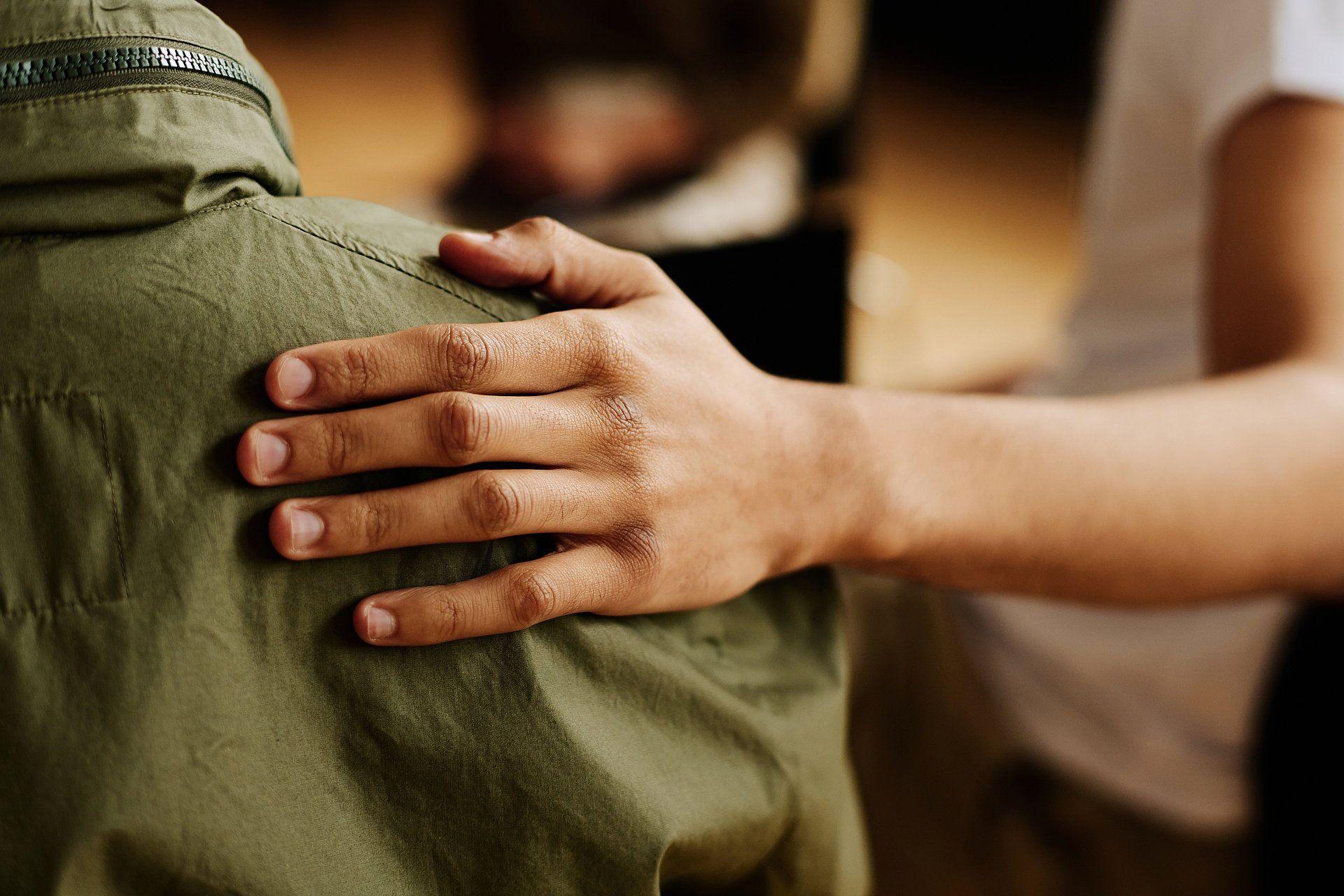 Eine unterstützende Hand liegt auf der Schulter einer Person in einer grünen Jacke – ein Symbol für Mitgefühl, Unterstützung und Zusammenhalt.
