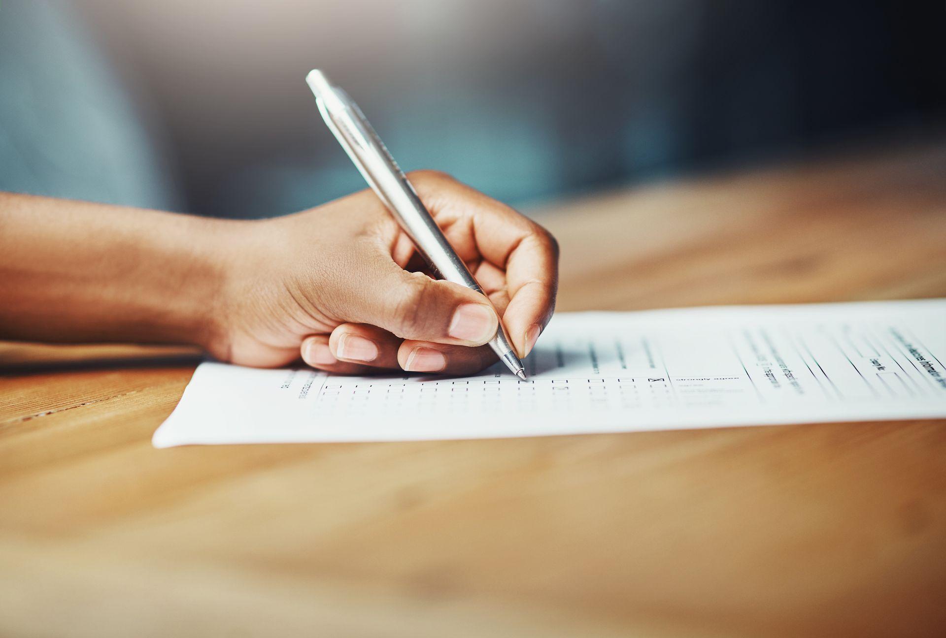 Eine Hand liegt auf einem Tisch und hält einen Kugelschreiber in der Hand. Darunter liegt ein Blatt Papier, welches die Person gerade ausfüllt. 