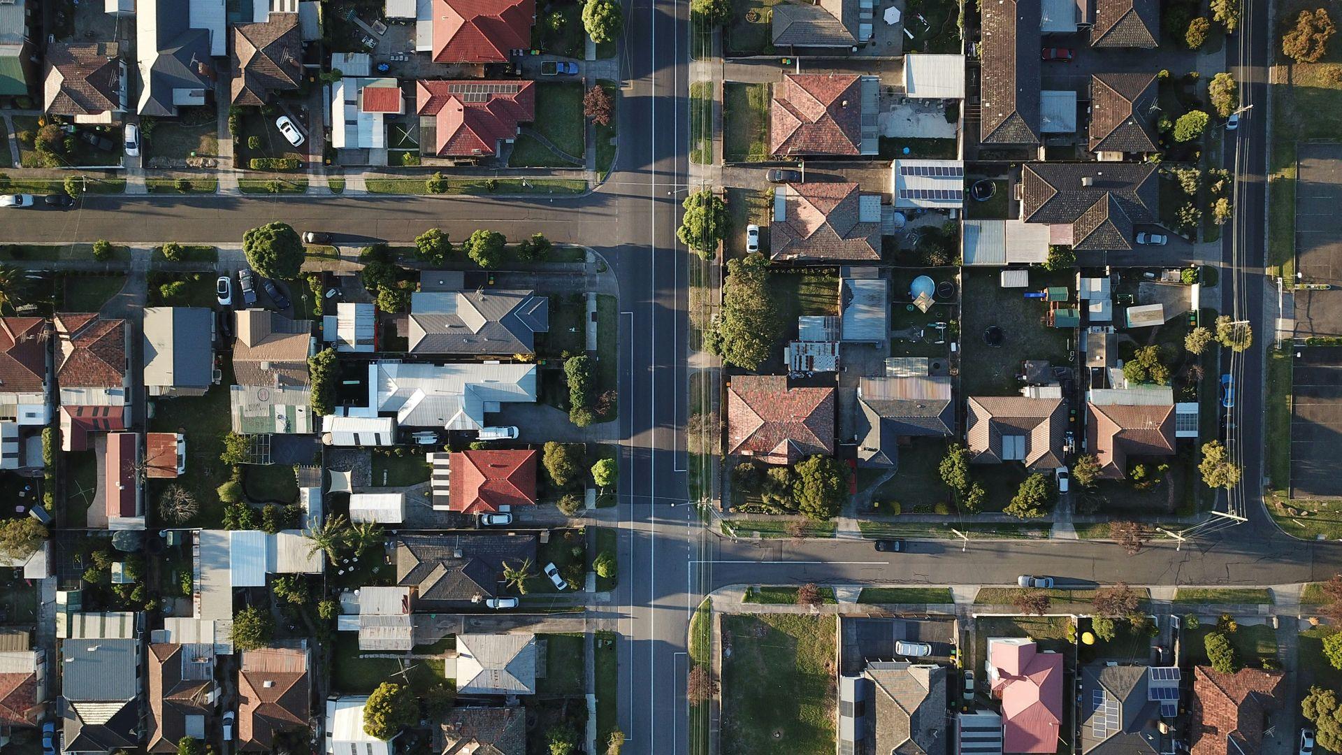 Luftaufnahme eines Wohnviertels mit zahlreichen Einfamilienhäusern, Gärten und Straßenkreuzungen. Die Dächer der Häuser sind in verschiedenen Farben gehalten, einige mit Solaranlagen ausgestattet.