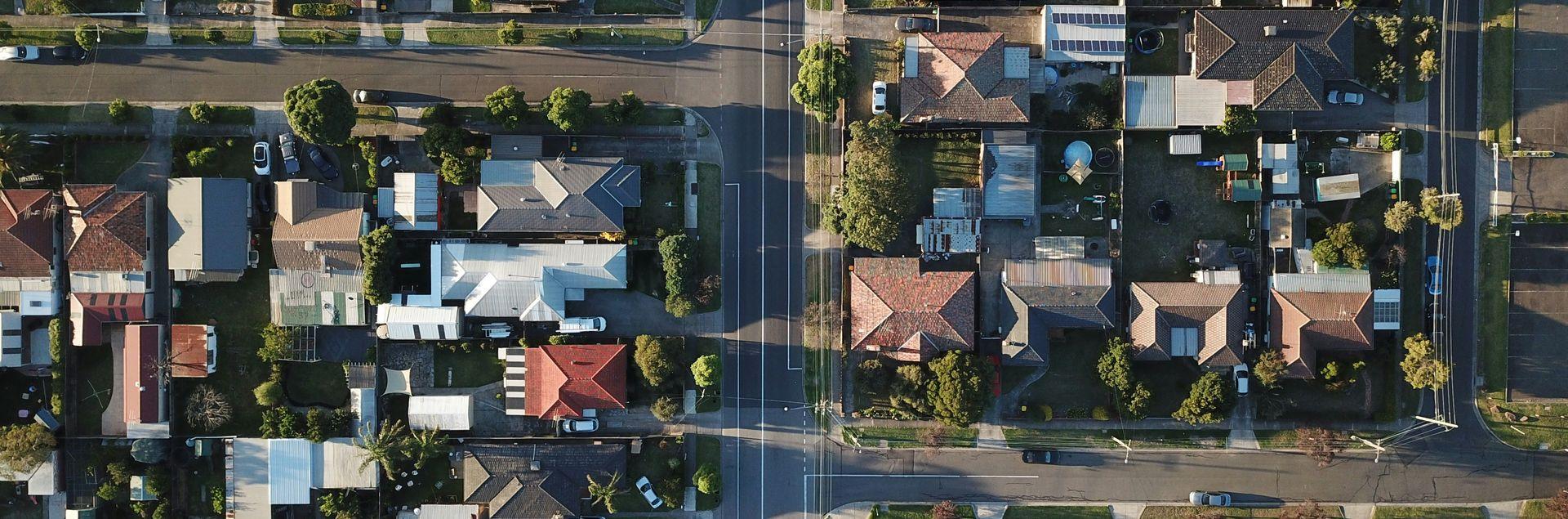 Luftaufnahme eines Wohnviertels mit zahlreichen Einfamilienhäusern, Gärten und Straßenkreuzungen. Die Dächer der Häuser sind in verschiedenen Farben gehalten, einige mit Solaranlagen ausgestattet.