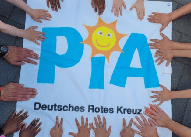 Mehrere Kinder- und Erwachsenenhände legen sich rund um ein Banner mit der Aufschrift „PIA“ und einem lachenden Sonnensymbol. Darunter steht „Deutsches Rotes Kreuz“. Mehrere Kinder- und Erwachsenenhände legen sich rund um ein Banner mit der Aufschrift „PIA“ und einem lachenden Sonnensymbol. Darunter steht „Deutsches Rotes Kreuz“.