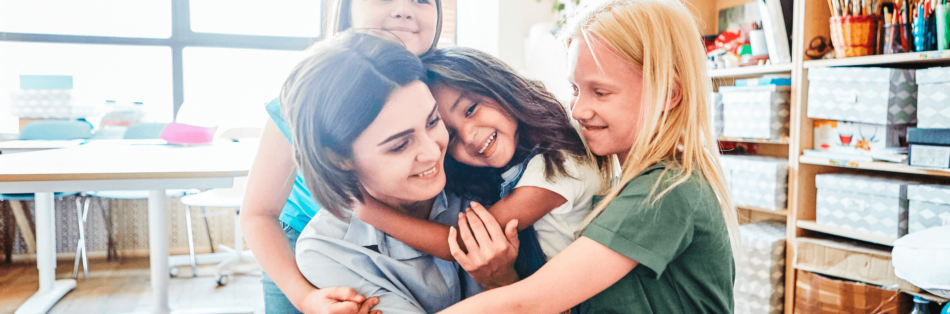 Eine lächelnde Frau wird in einem kreativen Raum herzlich von drei fröhlichen Kindern umarmt. Eine lächelnde Frau wird in einem kreativen Raum herzlich von drei fröhlichen Kindern umarmt.