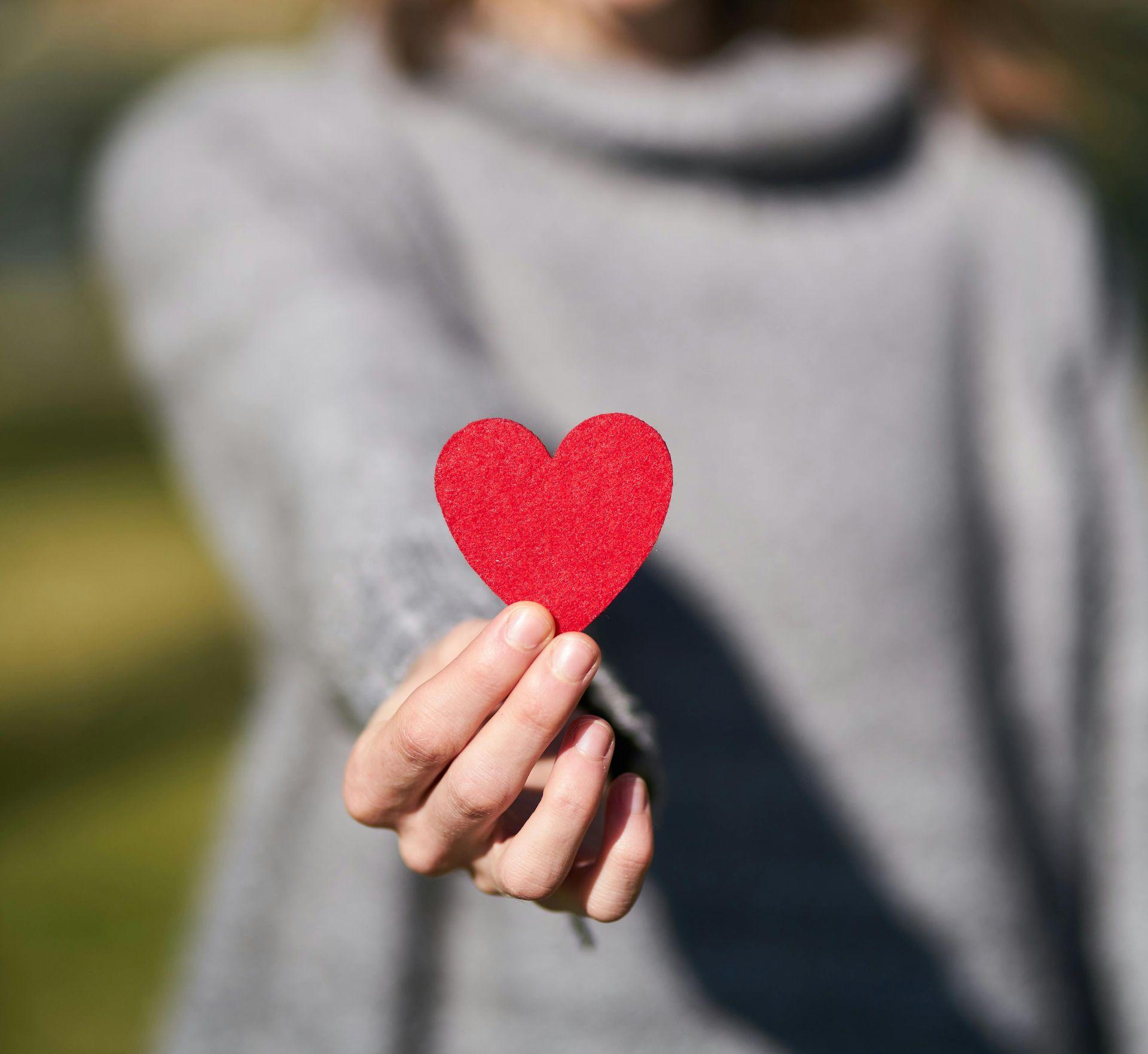 Eine Person hält ein rotes Herz aus Filz in die Kamera, während ihr Gesicht unscharf im Hintergrund bleibt. Eine Person hält ein rotes Herz aus Filz in die Kamera, während ihr Gesicht unscharf im Hintergrund bleibt.