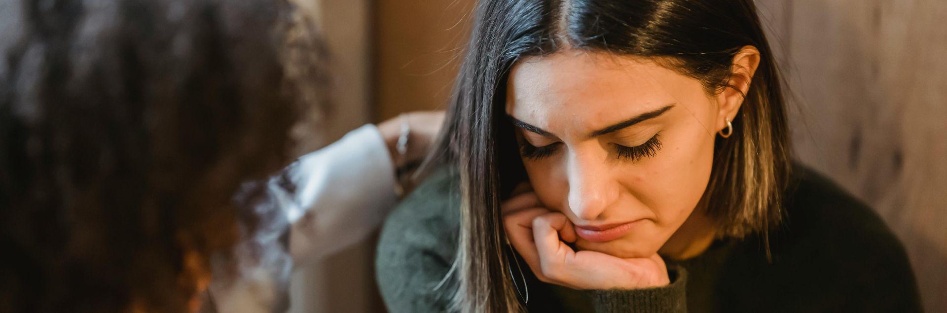 Eine junge Frau schaut bedrückt auf den Boden und eine andere versucht diese zu trösten Eine junge Frau schaut bedrückt auf den Boden und eine andere versucht diese zu trösten