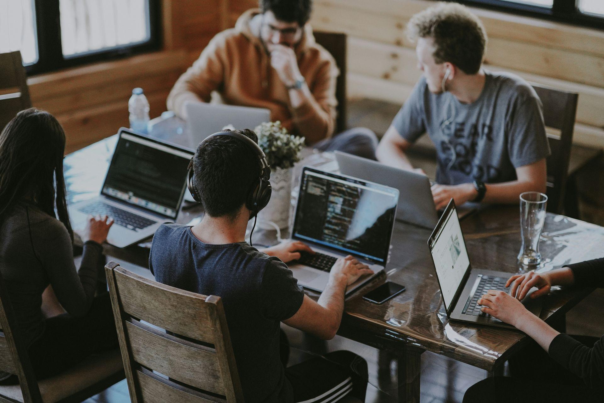 Eine Gruppe junger Menschen sitzt um einen Tisch herum, alle mit Laptops beschäftigt. Einer trägt Kopfhörer, andere diskutieren oder konzentrieren sich auf ihre Bildschirme. Die Umgebung wirkt gemütlich und hat einen hölzernen Innenraum, was eine entspannte Arbeitsatmosphäre vermittelt. Eine Gruppe junger Menschen sitzt um einen Tisch herum, alle mit Laptops beschäftigt. Einer trägt Kopfhörer, andere diskutieren oder konzentrieren sich auf ihre Bildschirme. Die Umgebung wirkt gemütlich und hat einen hölzernen Innenraum, was eine entspannte Arbeitsatmosphäre vermittelt.