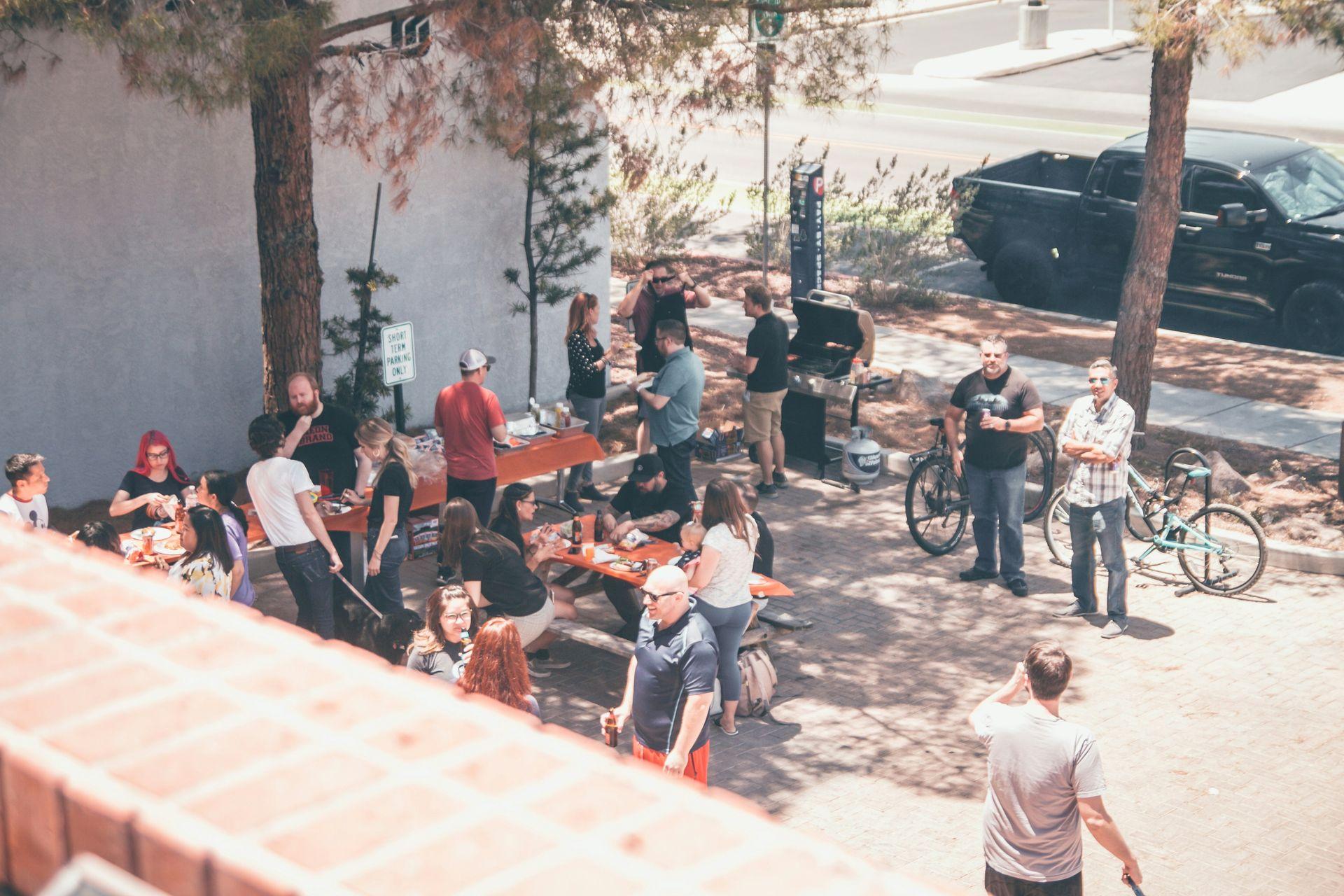 Eine Gruppe von Menschen trifft sich im Freien zu einem Grillfest mit Bänken, Essen und Getränken. Einige unterhalten sich, während andere am Grill stehen oder herumlaufen. Fahrräder und ein schwarzer Pickup sind im Hintergrund zu sehen. Eine Gruppe von Menschen trifft sich im Freien zu einem Grillfest mit Bänken, Essen und Getränken. Einige unterhalten sich, während andere am Grill stehen oder herumlaufen. Fahrräder und ein schwarzer Pickup sind im Hintergrund zu sehen.
